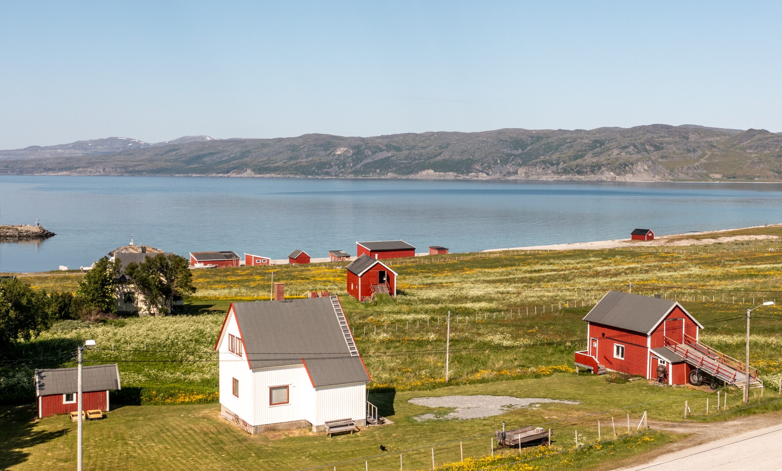 Nillagården, hvitt bolighus, rød fjøs og uthus. I bakgrunnen en annen gård, en rekke røde naust og fjord og fjell. Fint vær