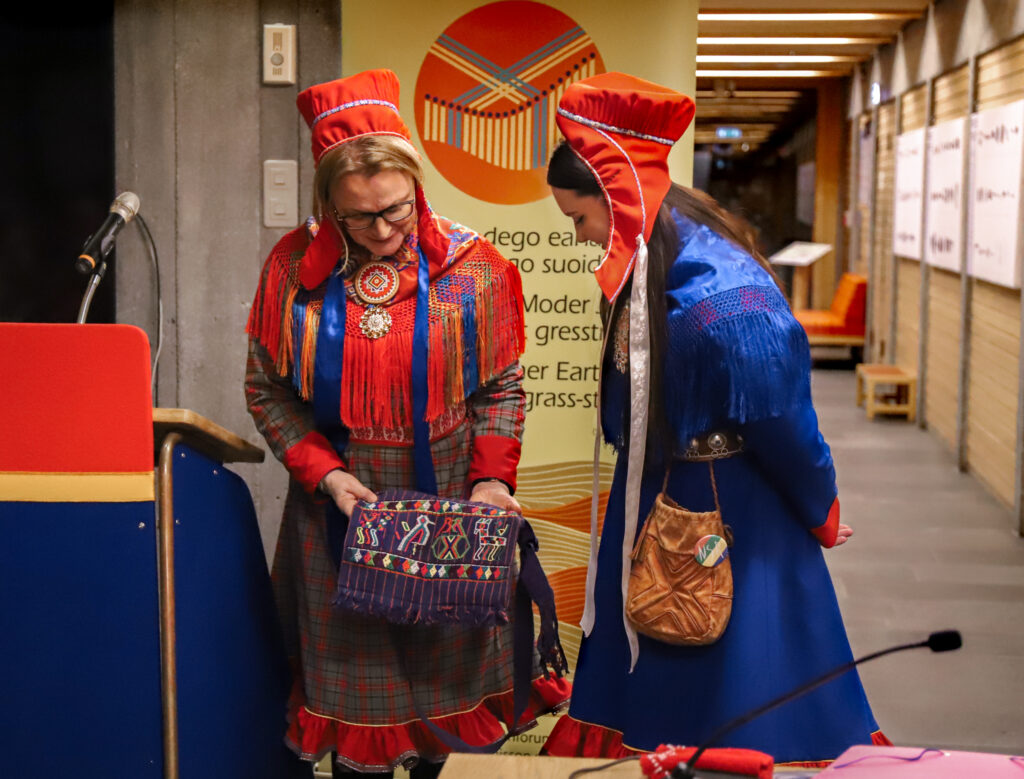 Two Sámi women in traditional clothing admiring a woven bag.