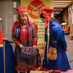 Two Sámi women in traditional clothing admiring a woven bag.