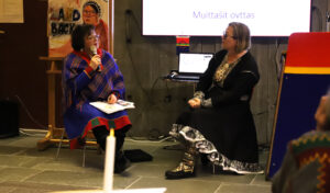 Two Sámi women in traditional clothing sitting and discussing.